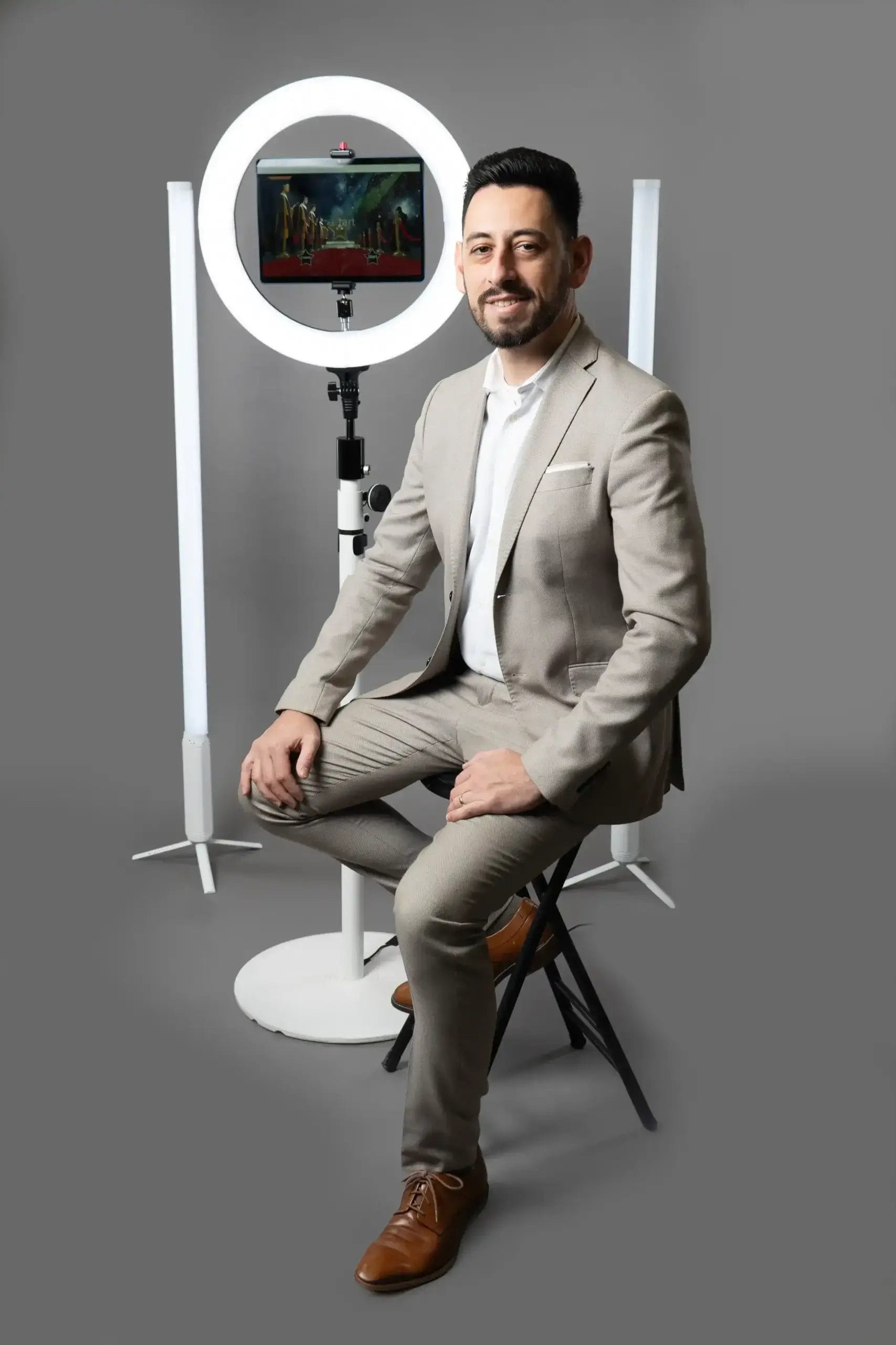 A man in a beige suit sits on a stool, smiling at the camera. Behind him are a circular ring light, a tablet on a stand, and two vertical LED lights, all set against a plain gray background.