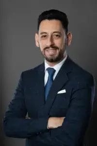A man with short dark hair and a beard, wearing a blue checked suit, white shirt, and blue tie, stands with arms crossed and smiles slightly against a plain gray background.