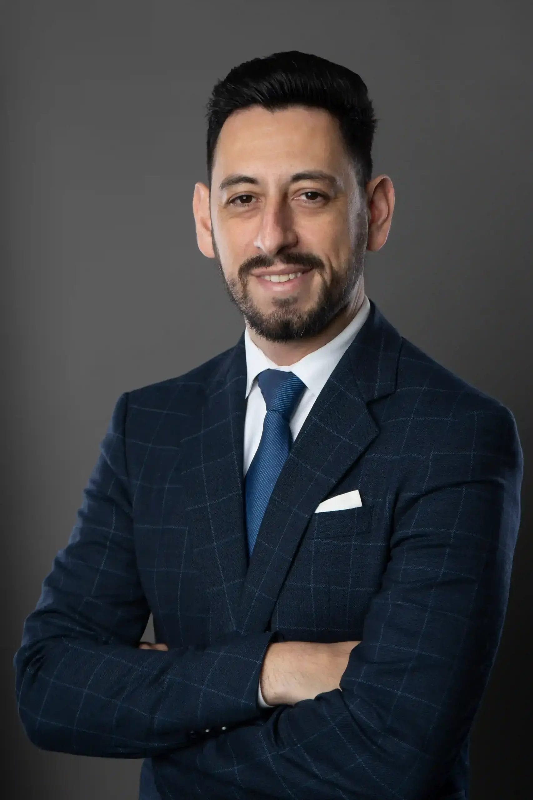 A man with short dark hair and a beard, wearing a blue checked suit, white shirt, and blue tie, stands with arms crossed and smiles slightly against a plain gray background.
