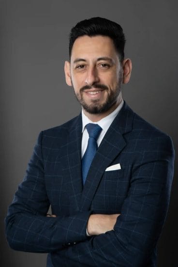 A man with short dark hair and a beard, wearing a blue checked suit, white shirt, and blue tie, stands with arms crossed and smiles slightly against a plain gray background.