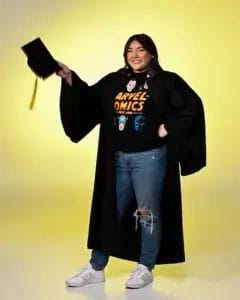A young person in a graduation gown and Marvel Comics T-shirt smiles and holds up a graduation cap. They are wearing ripped jeans and white sneakers, standing against a yellow background.