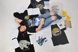 A smiling person with purple hair lies on the floor surrounded by choir T-shirts, sheet music, and awards, holding a choir music binder and wearing a black "Region Choir" T-shirt and ripped blue jeans.