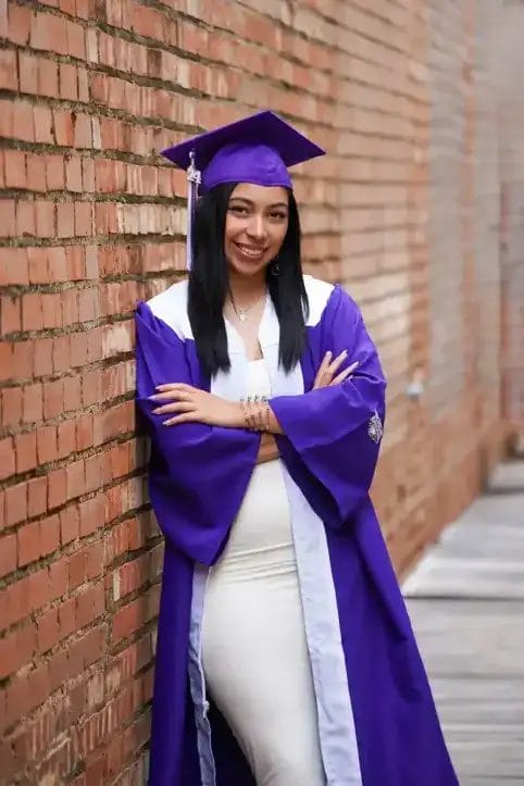 A young woman in a purple graduation cap and gown stands smiling with arms crossed, leaning against a brick wall on a wooden walkway.