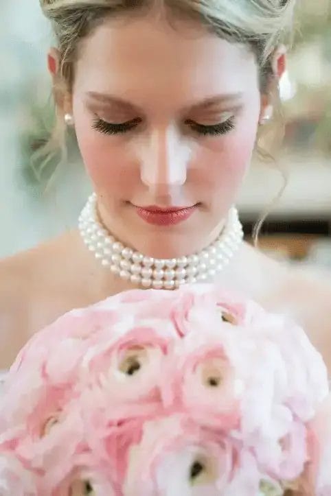 A bride with blonde hair and pearl jewelry looks down at a bouquet of light pink flowers, her eyes closed, with soft makeup and a serene expression.