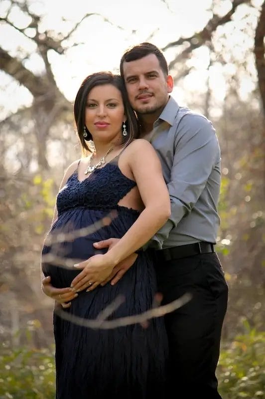 A pregnant woman in a dark dress stands outdoors, holding her belly, while a man in a grey shirt stands closely behind her with his arms around her. Both look at the camera, surrounded by trees in the background.