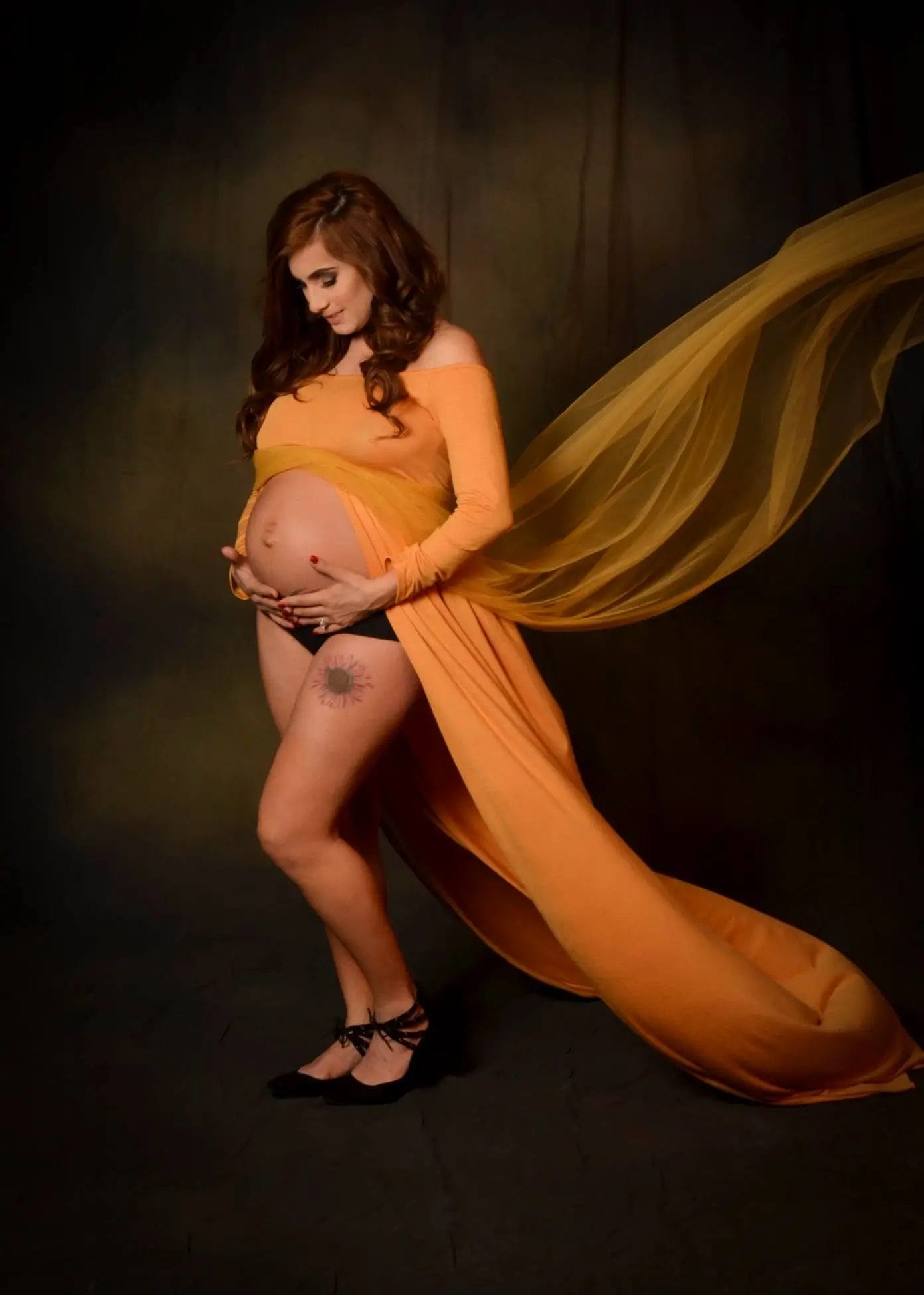 A pregnant woman in a flowing orange gown cradles her belly, with sheer fabric billowing behind her. She stands barefoot on dark flooring against a dark backdrop, revealing a tattoo on her thigh.