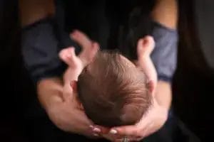 A close-up view from above of a woman cradling a newborn baby in her hands, focusing on the back of the baby's head and tiny feet. The woman's face is out of focus in the background.