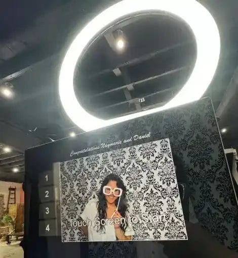 A touchscreen photo booth displays "Congratulations Naynarie and Daniel" above a photo of a smiling woman in oversized glasses, with a ring light and patterned wallpaper background. "Touch Screen to Start!" is shown on the screen.