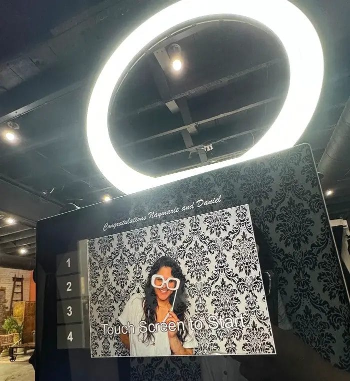 A photo booth screen displays a woman with long hair and oversized glasses posing in front of a patterned backdrop. Above the screen, a bright ring light glows. The text says "Congratulations Naymarie and Daniel.