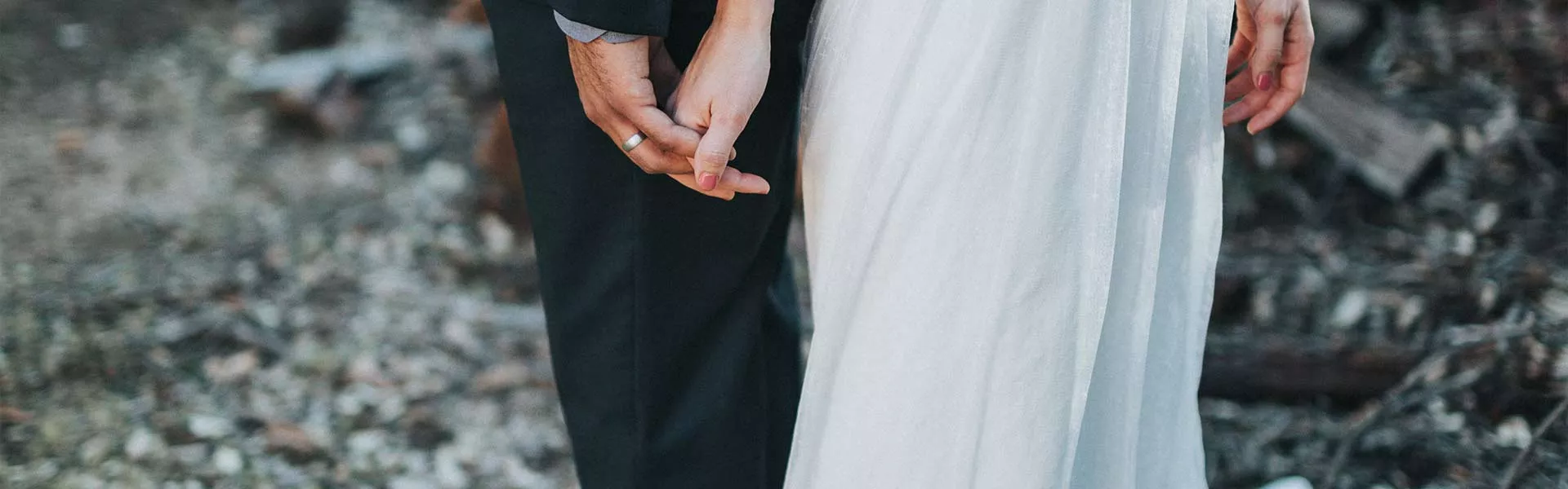 Close-up of a couple holding hands, with one person wearing a black suit and the other wearing a white wedding dress, standing outdoors on a rocky ground.