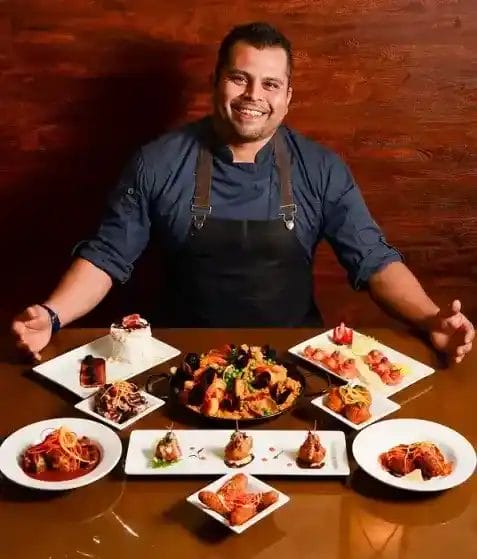 A smiling chef in a dark apron sits at a table displaying a variety of colorful, plated dishes including seafood, pasta, sushi, desserts, and appetizers in front of a brown wooden background.