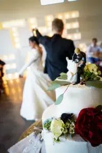 A wedding cake with floral decorations and a bride-and-groom topper is in focus, while a bride and groom dance in the blurred background. Warm lights and guests can be seen in the softly lit reception hall.