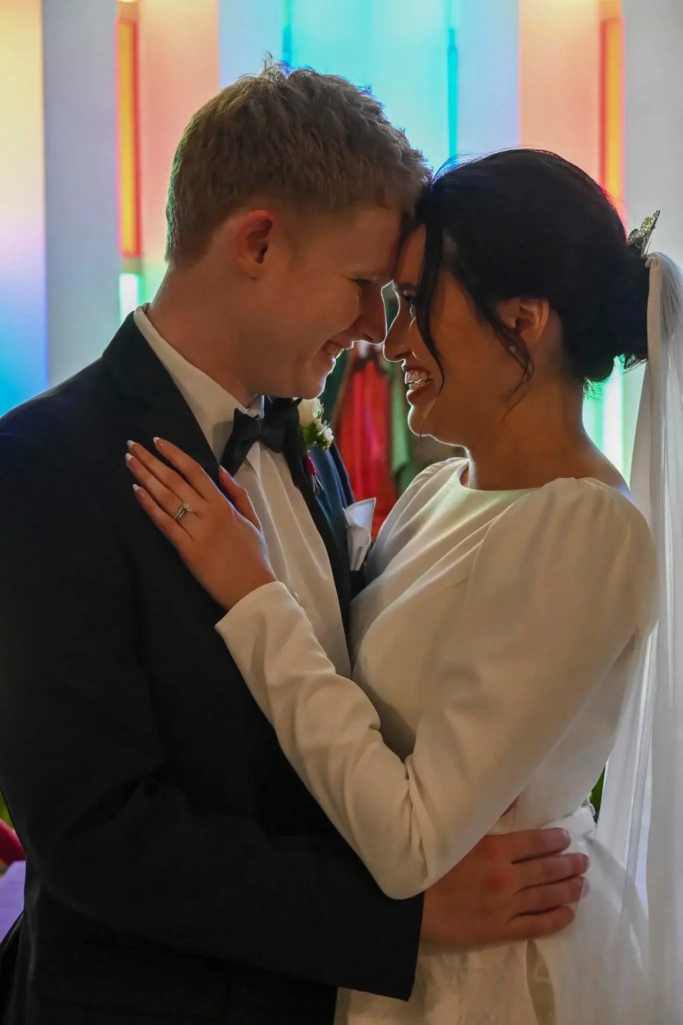 A bride and groom in wedding attire embrace and smile, touching foreheads in front of colorful stained glass windows.