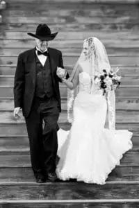 A bride in a wedding gown holding a bouquet walks down wooden steps, holding hands with an older man in a dark suit and cowboy hat. Both appear happy and focused ahead. The image is black and white.