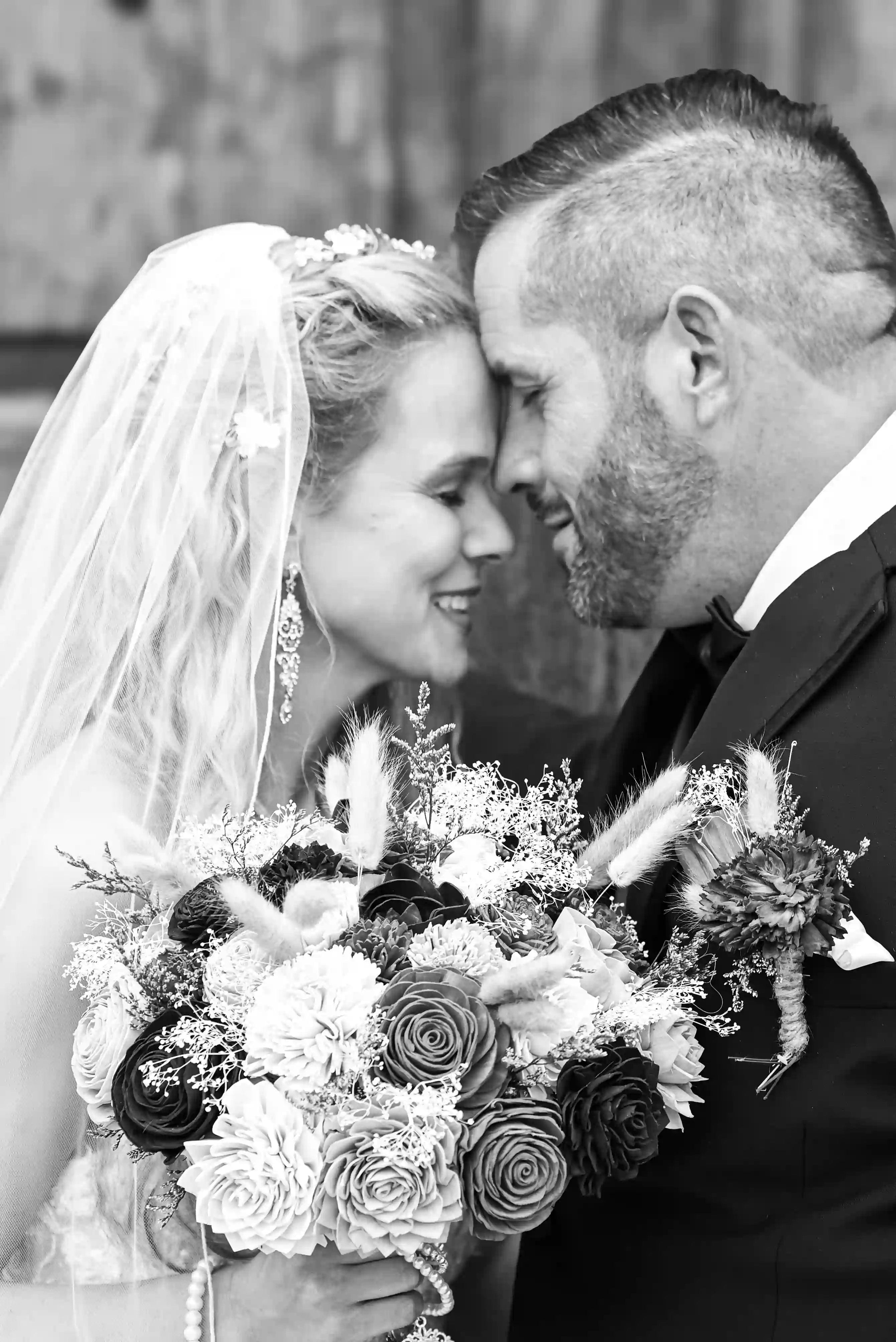 A bride and groom stand close together, smiling with their foreheads touching. The bride holds a large bouquet of flowers and wears a veil, while the groom is in a suit. The image is in black and white.