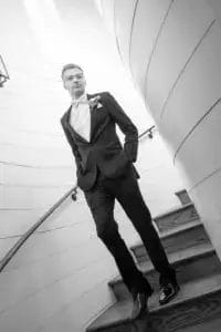 A young man in a tuxedo walks down a spiral wooden staircase, hands in pockets, looking confident. The scene is in black and white with soft, natural lighting from above.