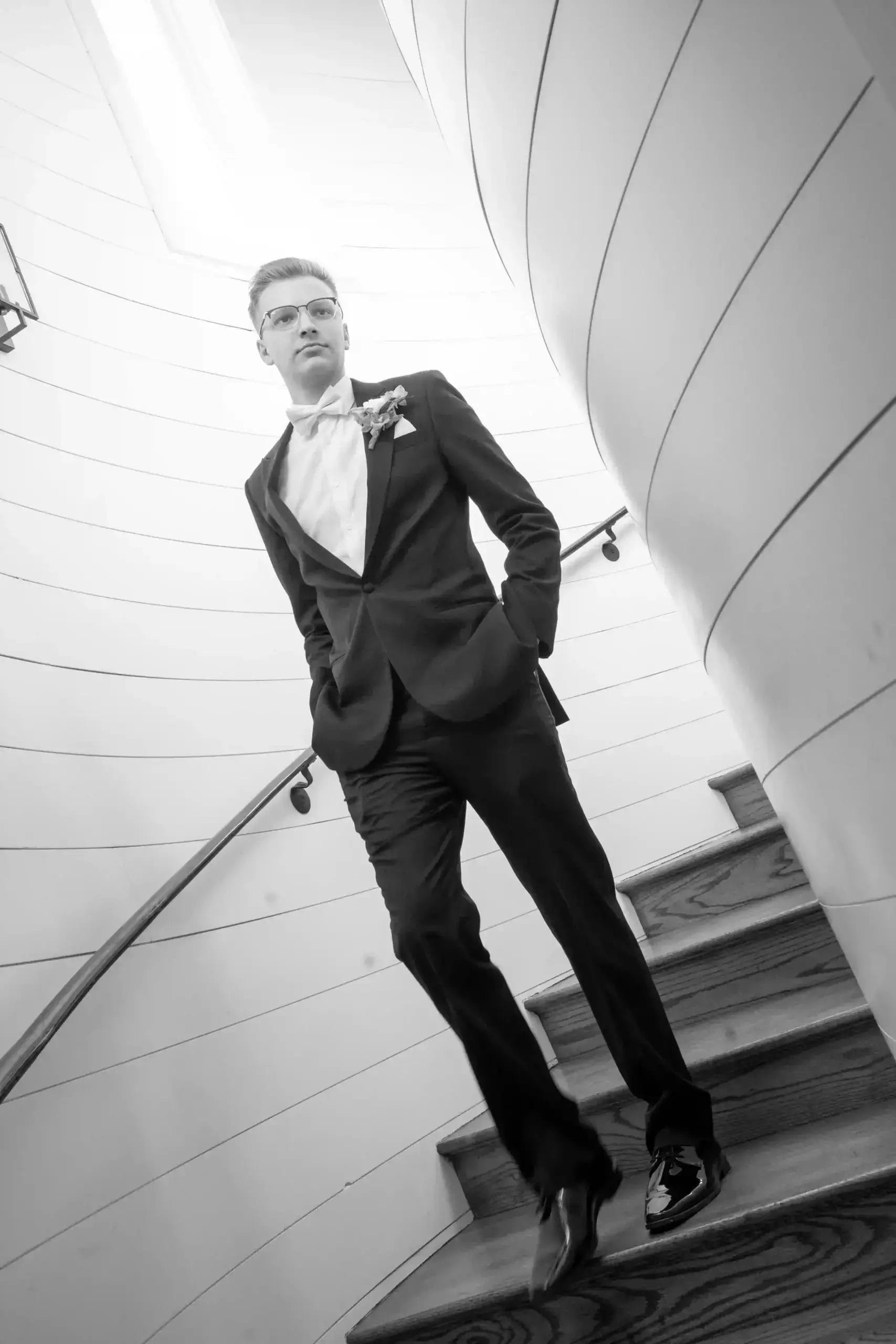 A young man in a tuxedo walks down a spiral wooden staircase, hands in pockets, looking confident. The scene is in black and white with soft, natural lighting from above.