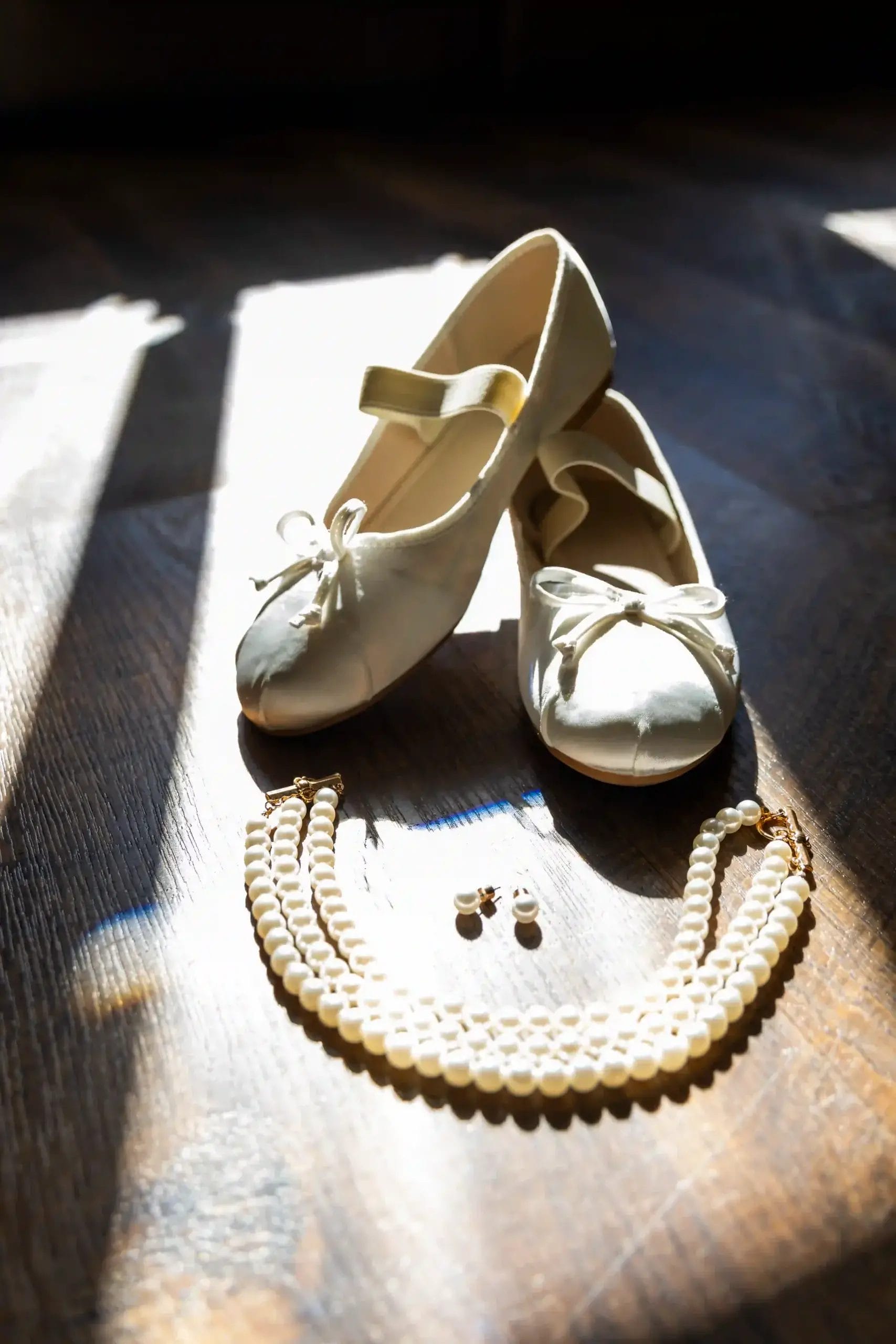 A pair of white ballet flats with bows, a double-strand pearl necklace, and pearl earrings are arranged on a wooden floor in bright sunlight.