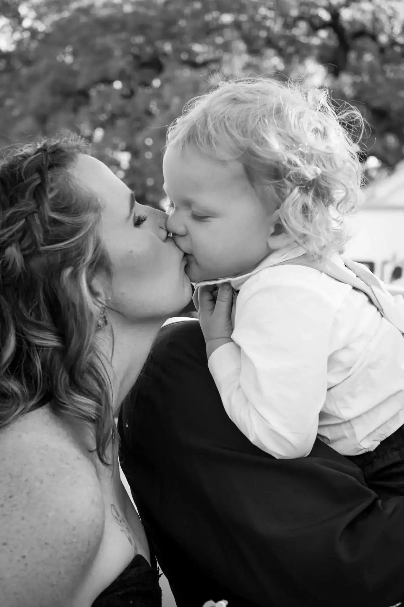 A woman gently kisses a young child on the lips while holding them close. The child has curly hair and wears a light shirt. The photo is in black and white, and they are outdoors with trees in the background.