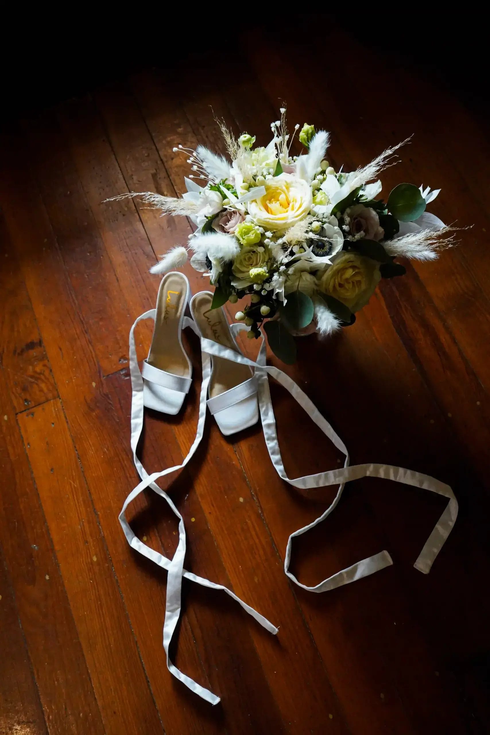 A bouquet of white and yellow flowers rests on a wooden floor next to a pair of white, open-toe bridal shoes with long ribbon straps.