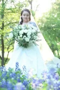 A bride in a white dress and veil stands outdoors, holding a large bouquet of white roses and greenery, with blurred purple flowers in the foreground and lush green trees in the background.