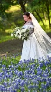 A bride in a white dress and long veil holds a large bouquet of white flowers, walking through a field of blooming bluebonnets with trees and sunlight in the background.