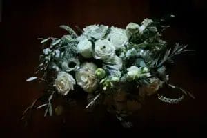 A bouquet of white roses and greenery is arranged in a loose, natural style against a dark background, with soft lighting highlighting the delicate petals and leaves.