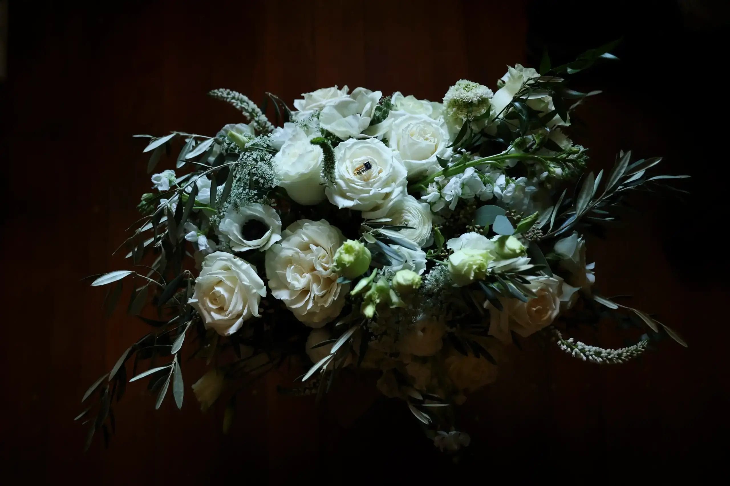 A bouquet of white roses and greenery is arranged in a loose, natural style against a dark background, with soft lighting highlighting the delicate petals and leaves.