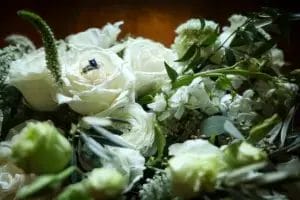 A blue gemstone ring nestled inside a white rose, surrounded by various white flowers and green foliage, softly lit in a warm, intimate setting.