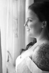 A woman in a wedding dress with floral tattoos on her shoulder smiles as she looks out a window, softly lit by natural light through sheer curtains. The image is in black and white.