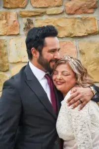A man in a suit and tie hugs a smiling woman in a white lace dress with a tiara. They stand close together in front of a stone wall, appearing happy and affectionate.