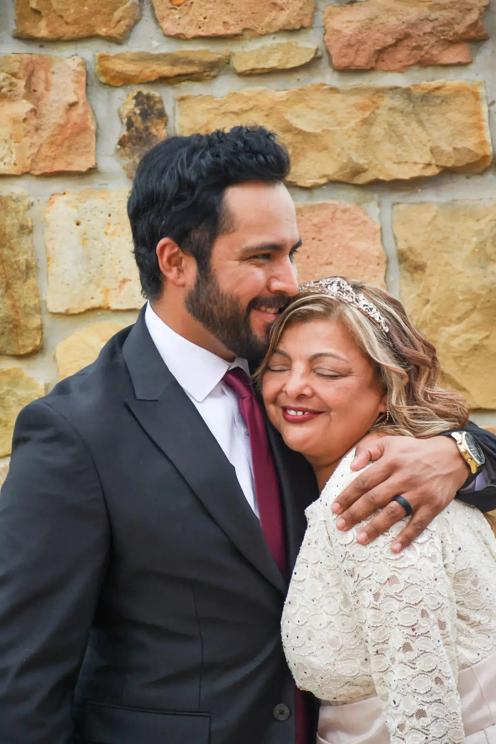 A man in a suit and tie hugs a smiling woman in a white lace dress with a tiara. They stand close together in front of a stone wall, appearing happy and affectionate.