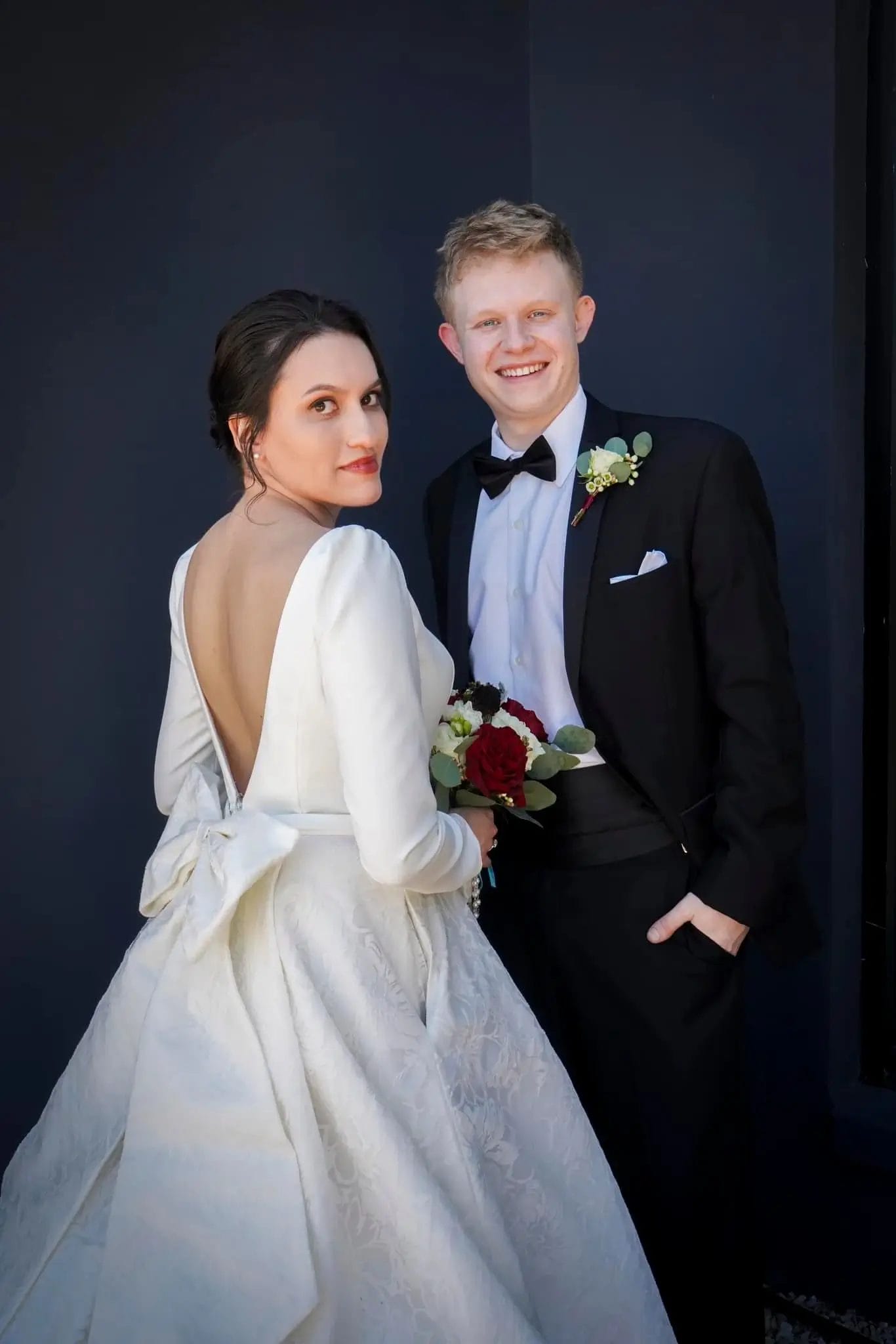 A bride in a white wedding dress with a low back and bow stands beside a groom in a black tuxedo. The bride holds a bouquet of red and white flowers; both are smiling against a dark background.