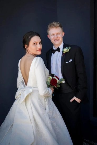 A bride in a white wedding dress with a low back and bow stands beside a groom in a black tuxedo. The bride holds a bouquet of red and white flowers; both are smiling against a dark background.