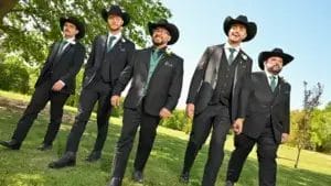 Five men dressed in black suits, green vests, and cowboy hats walk together smiling outdoors on a sunny day, with green grass and trees in the background.