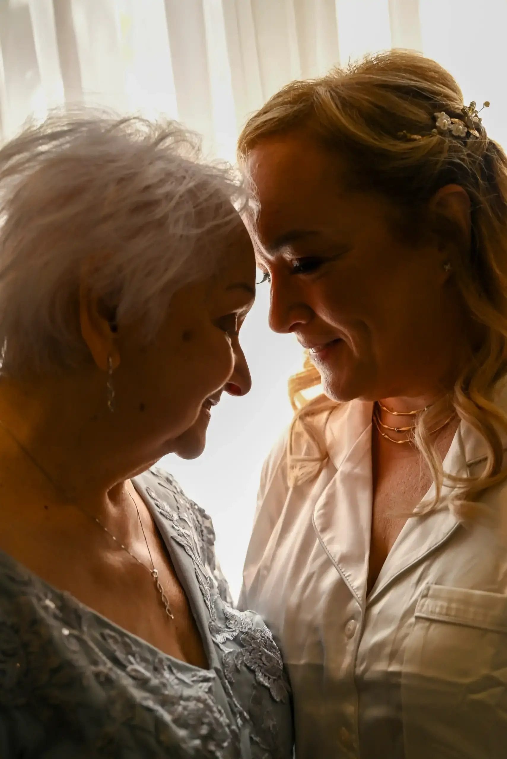 Two women, one older and one younger, stand close together indoors, smiling gently at each other with their foreheads touching, illuminated by soft light from a window behind them.