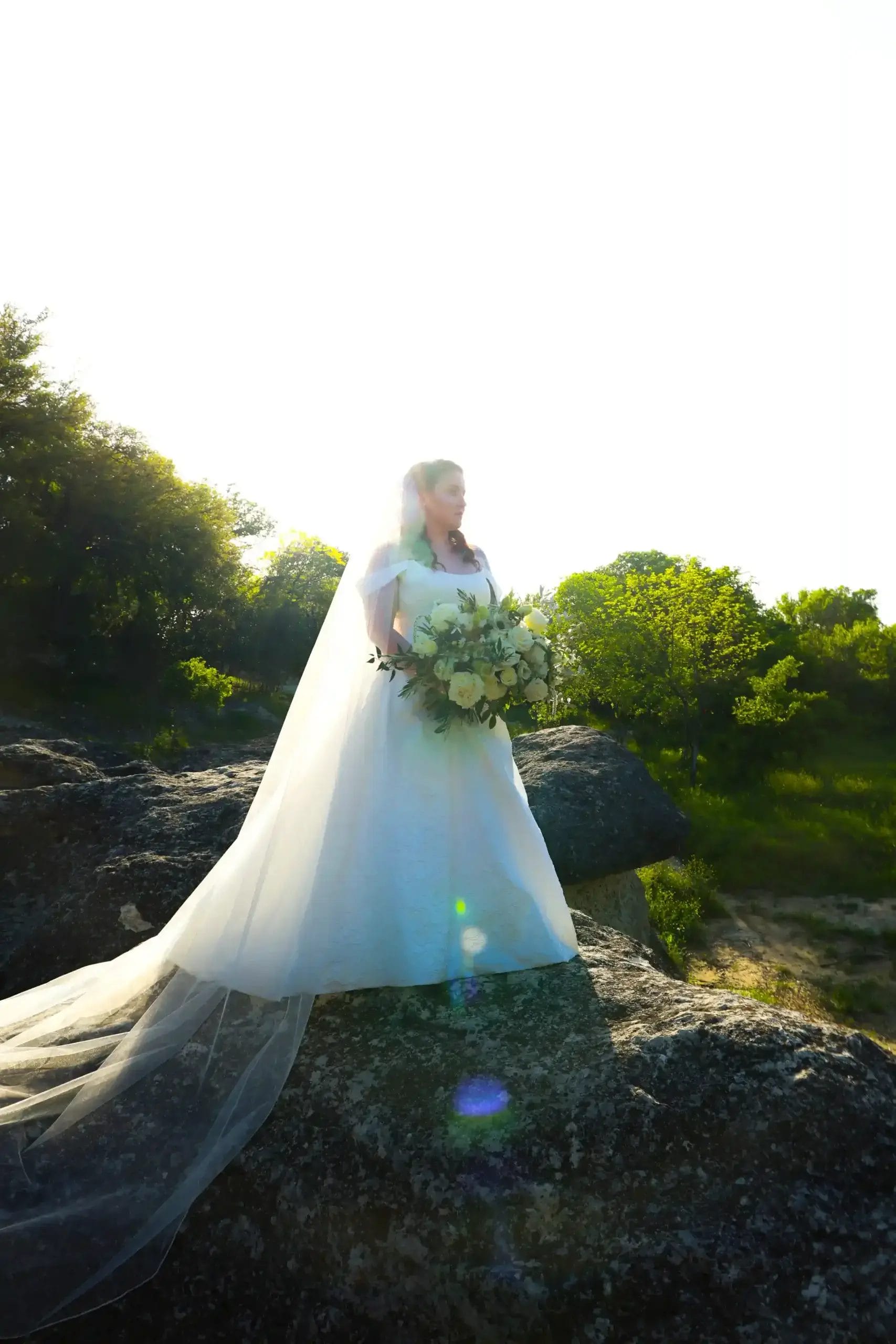 A bride in a white gown and long veil stands on a large rock outdoors, holding a bouquet of white flowers, with greenery and bright sunlight in the background.