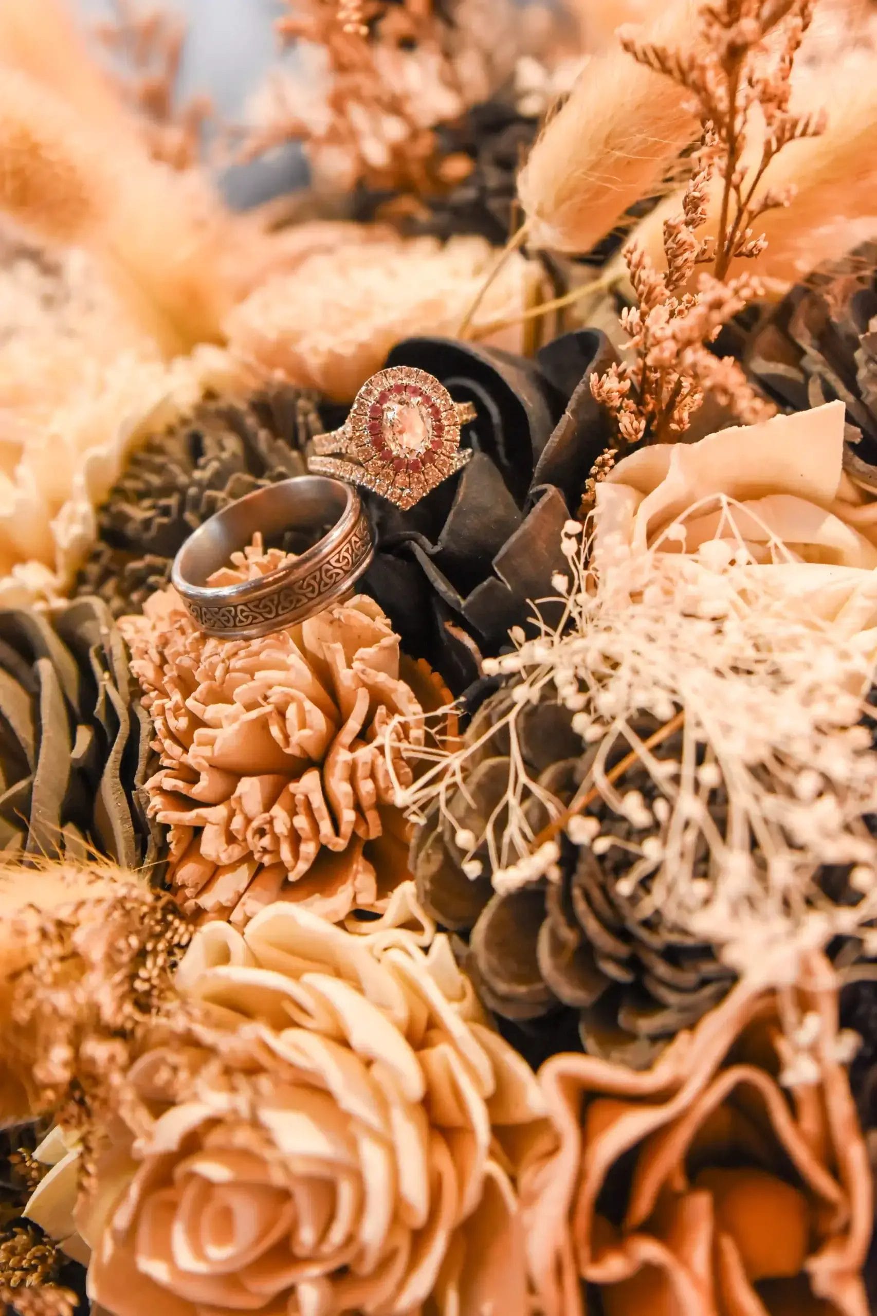 Two ornate rings rest on a bouquet of dried flowers and pinecones in warm beige and brown tones. The rings are surrounded by intricate textures from the floral arrangement.