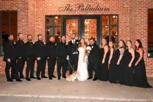 A wedding party poses in front of a brick building with “The Palladium” sign. The group includes six men in black suits, six women in black dresses, the bride in white, the groom in a tuxedo, and a small dog on a leash.