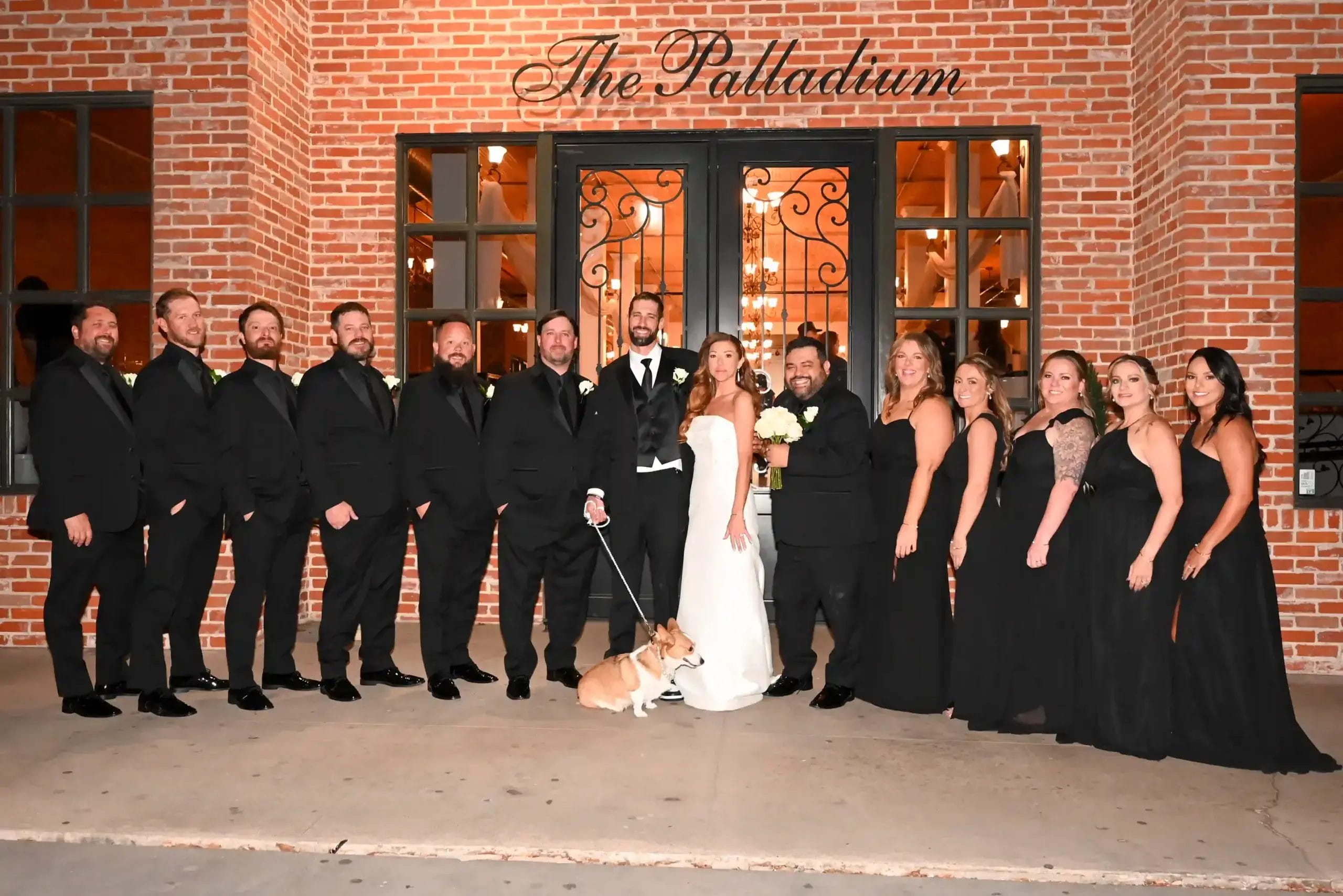 A wedding party poses in front of a brick building with “The Palladium” sign. The group includes six men in black suits, six women in black dresses, the bride in white, the groom in a tuxedo, and a small dog on a leash.