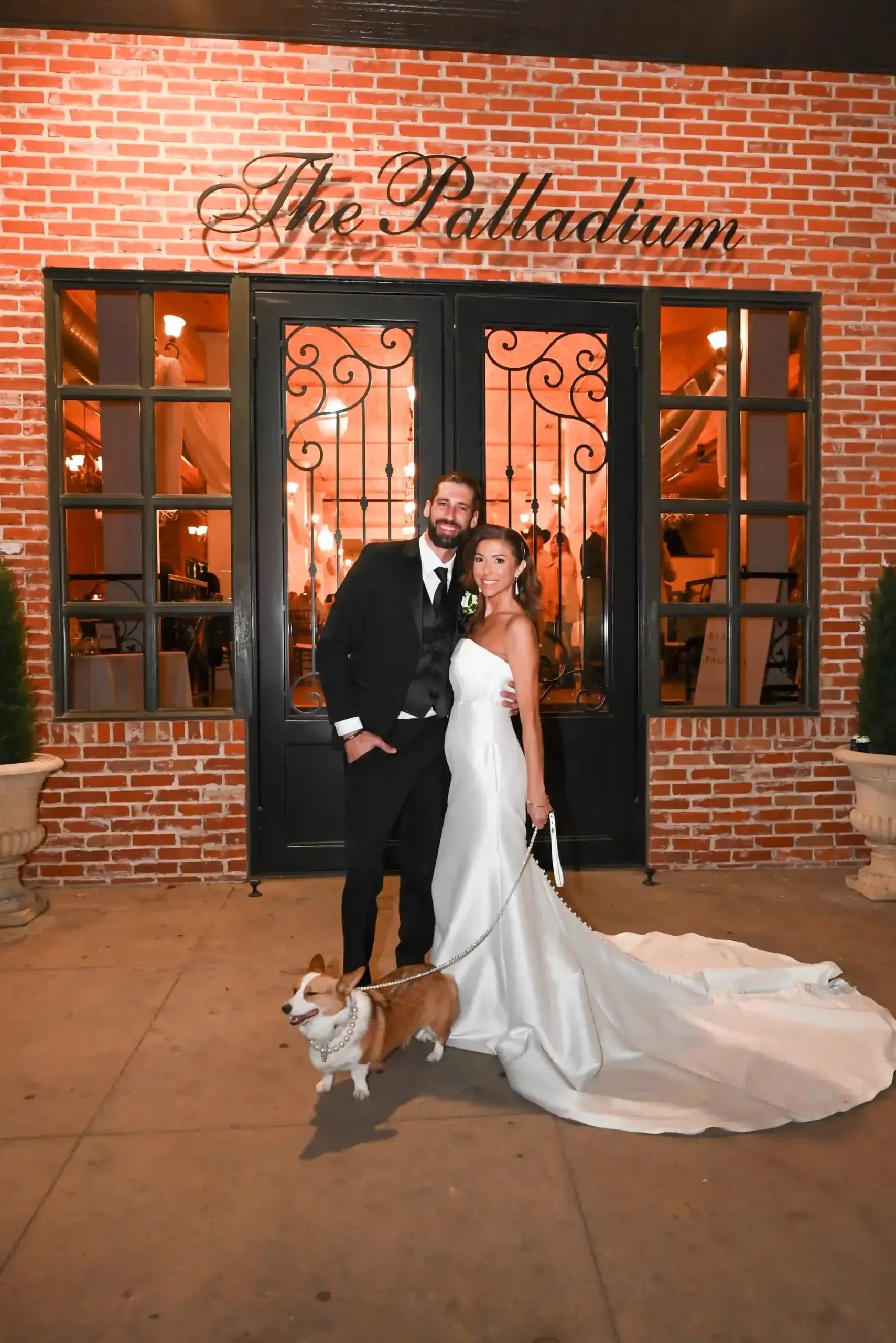 A bride and groom in formal wedding attire smile and pose outside a brick building labeled “The Palladium,” with their leashed Corgi dog standing in front of them. Warm lights glow indoors through large glass doors.