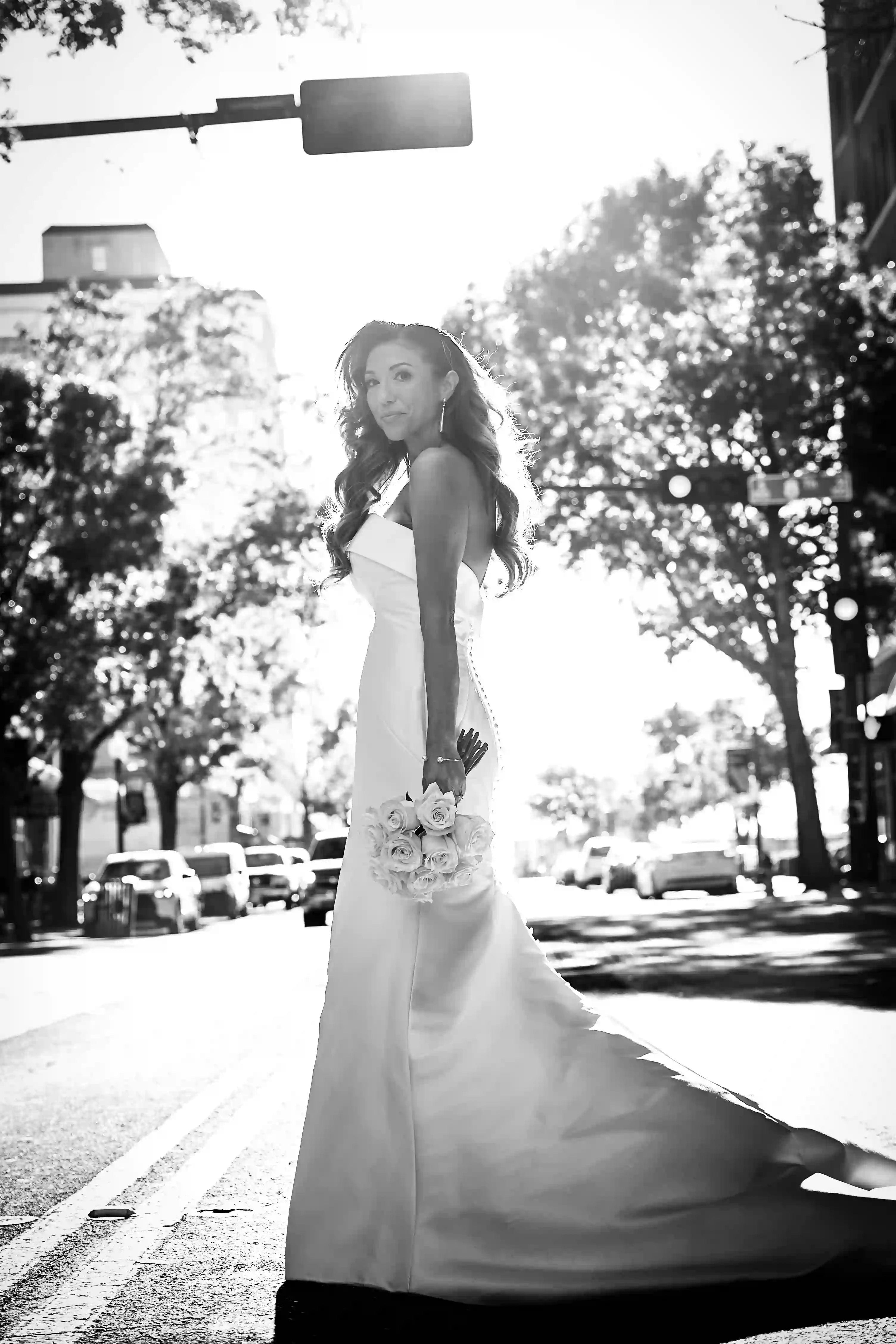 A bride in a long white gown holds a bouquet of flowers and smiles as she stands in the middle of a city street, sunlight streaming through the trees and buildings behind her.