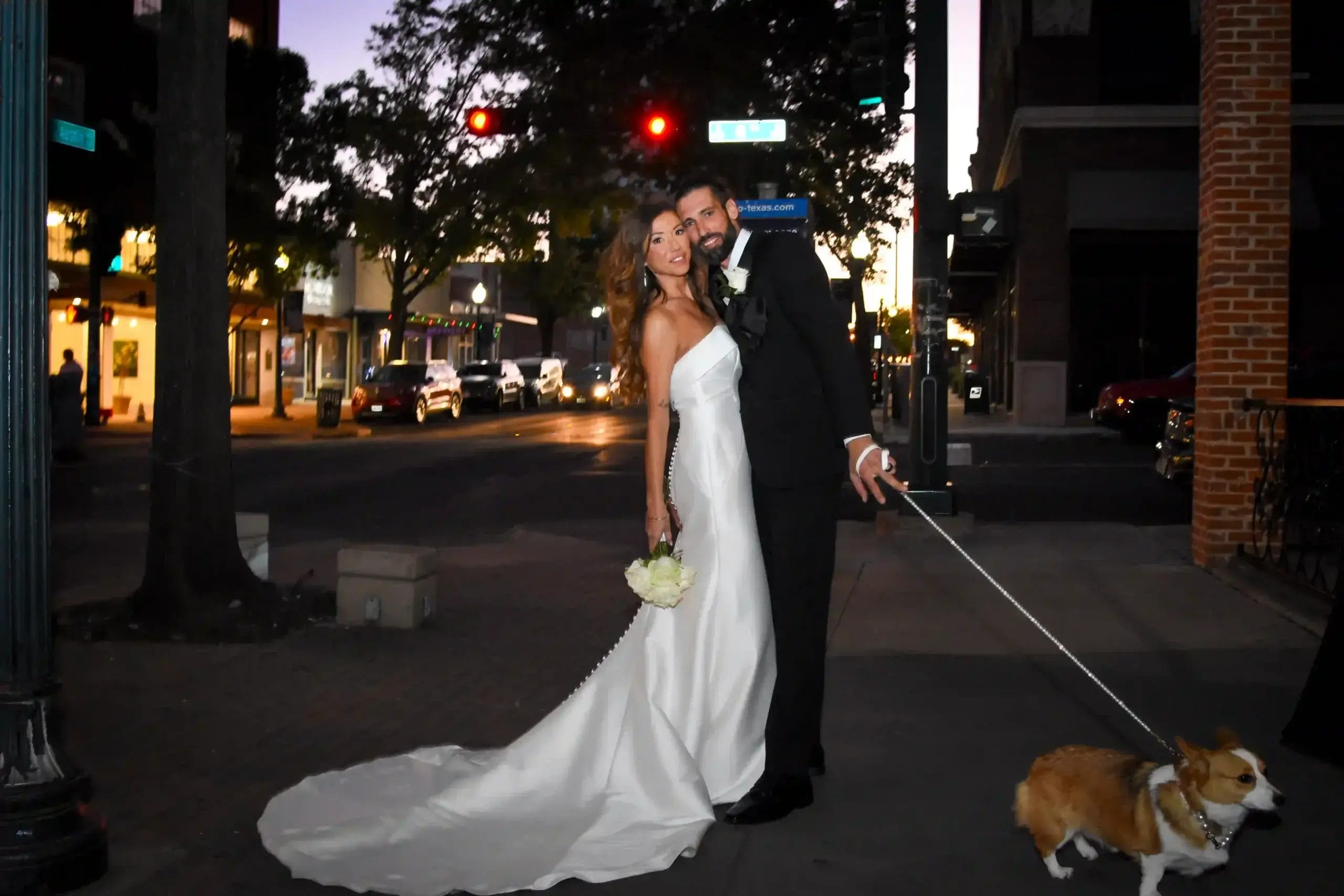 A bride in a white gown and a groom in a black suit smile and pose together on a city sidewalk at dusk, holding a bouquet and a corgi’s leash, with cars and streetlights in the background.