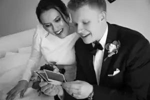 A bride and groom in wedding attire sit closely together on a couch, smiling as they look at several photographs. The bride holds a bouquet, and both appear joyful and relaxed.