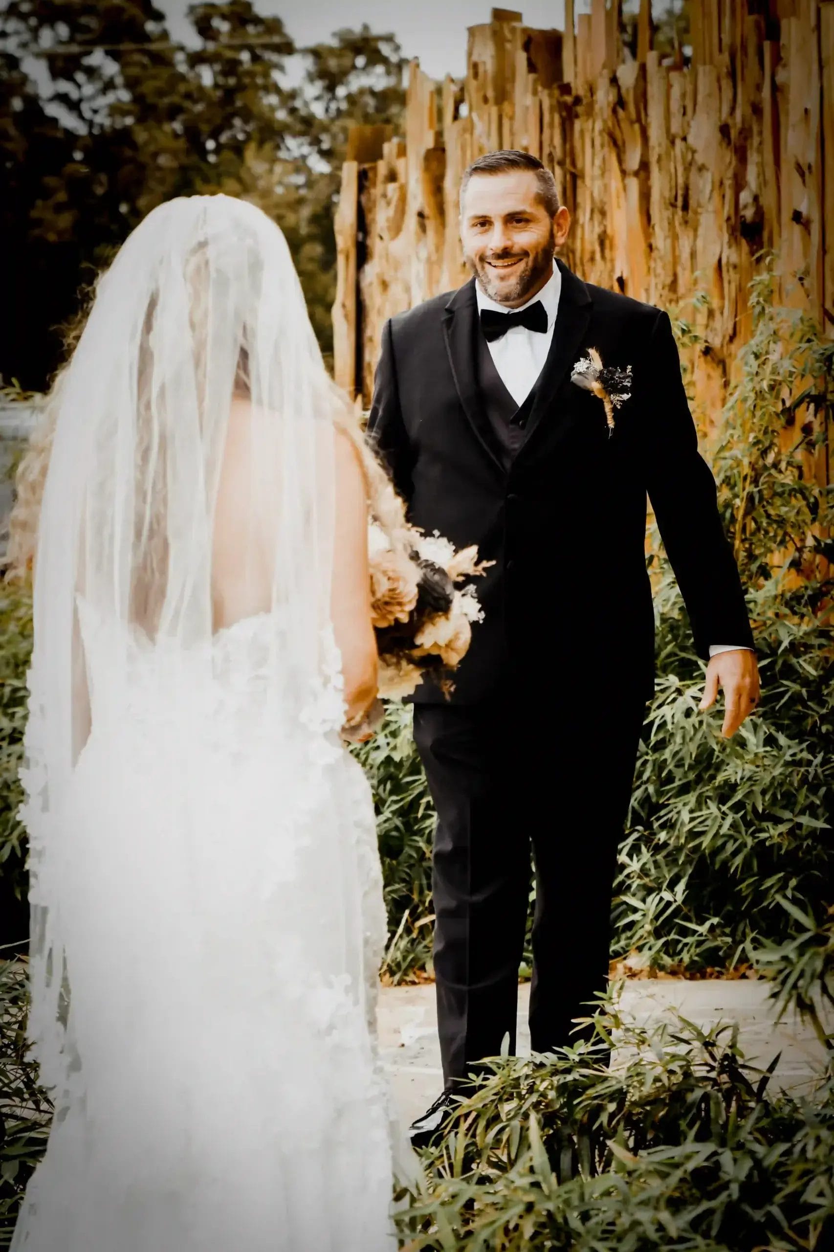 A bride in a white dress and veil stands facing a smiling groom in a black suit and bow tie, surrounded by greenery and a rustic wooden fence outdoors.