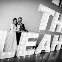 A bride and groom in wedding attire sit and smile beside large, illuminated marquee letters spelling “THE LEAH.” The photo is in black and white.