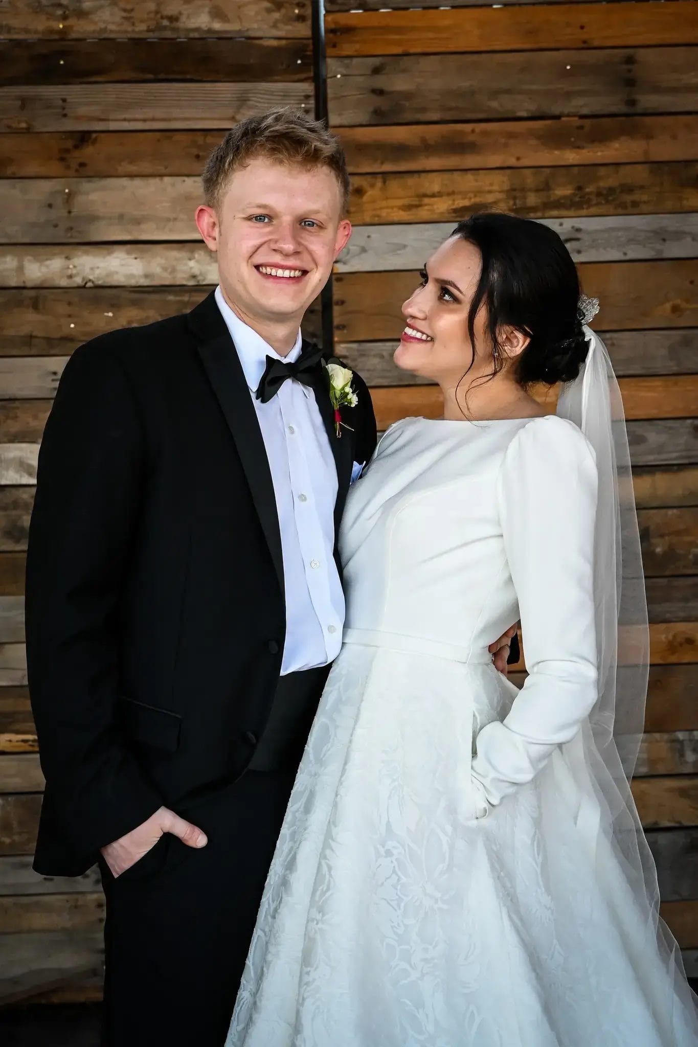 A smiling bride in a long-sleeved white gown and veil looks up at her groom, who is wearing a black tuxedo with a bow tie. They stand in front of a rustic wooden wall.