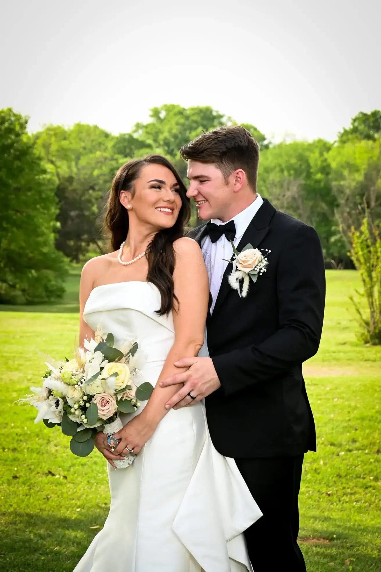 A bride in a white gown holding a bouquet smiles at her groom, who stands behind her in a black tuxedo. They are outdoors on a green lawn with trees in the background, looking happy and in love.