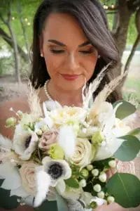 A bride with long brown hair and pearl necklace looks down, smiling softly, while holding a bouquet of white and pale pink flowers with green leaves and dried accents. Trees and greenery are blurred in the background.