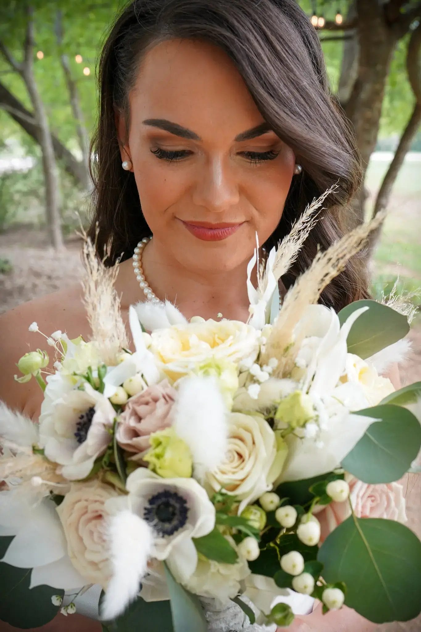 A bride with long brown hair and pearl necklace looks down, smiling softly, while holding a bouquet of white and pale pink flowers with green leaves and dried accents. Trees and greenery are blurred in the background.
